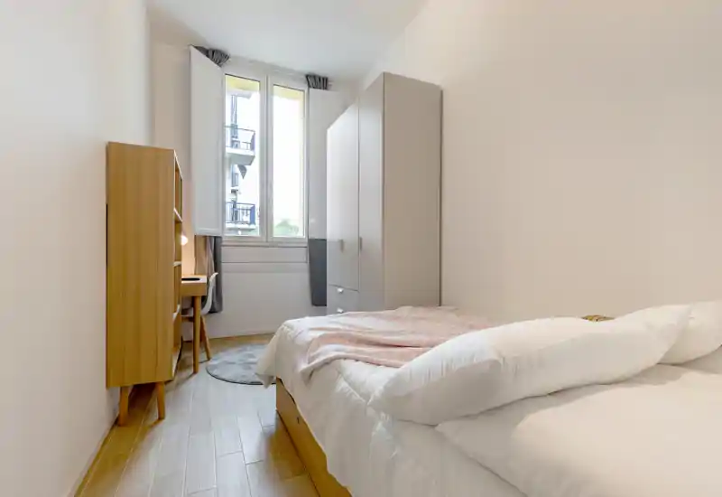 Dormitorio individual luminoso y ordenado con cama cómoda, armario y un pequeño escritorio junto a la ventana. La gran ventana aporta luz natural y la alfombra y la manta crean un ambiente acogedor.