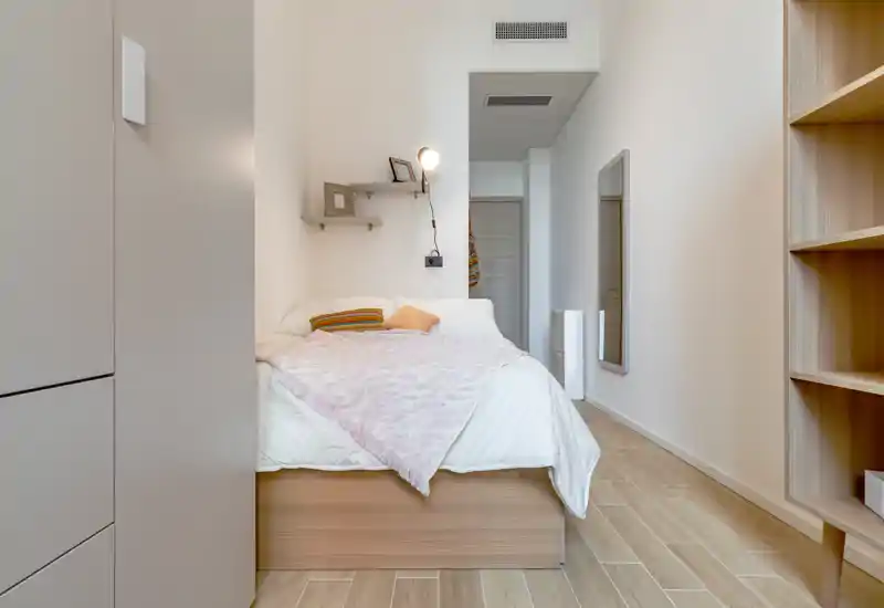 Dormitorio compacto y luminoso con cama arreglada, manta rosa y cojines decorativos; estanterías minimalistas y espejo de cuerpo entero amplían la sensación de espacio.