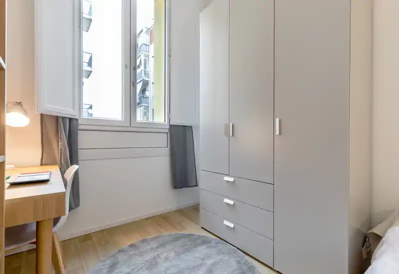 Rincón de dormitorio compacto con un gran armario, escritorio y ventana luminosa; tonos neutros y una alfombra redonda crean un área de descanso limpia y funcional.