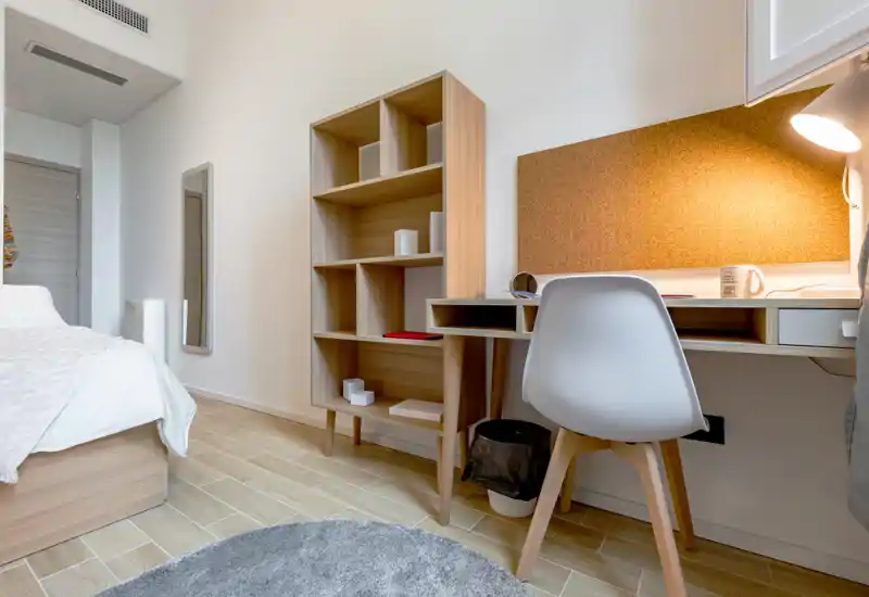 Dormitorio moderno y luminoso con cama baja de madera, escritorio con panel de corcho y lámpara, estantería abierta y alfombra suave — ideal para estudiante o profesional.