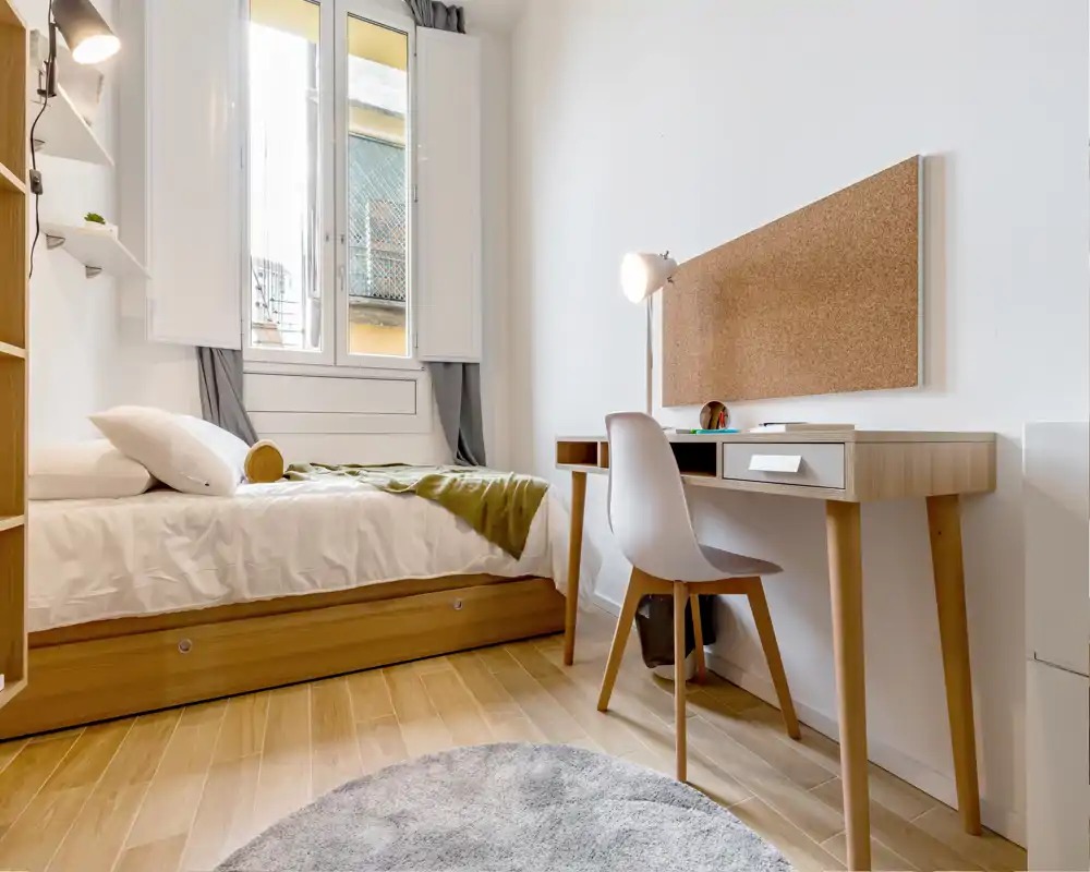 Dormitorio luminoso y compacto con cama individual, escritorio de madera y luz natural procedente de una gran ventana — ideal para estudiante o persona sola.