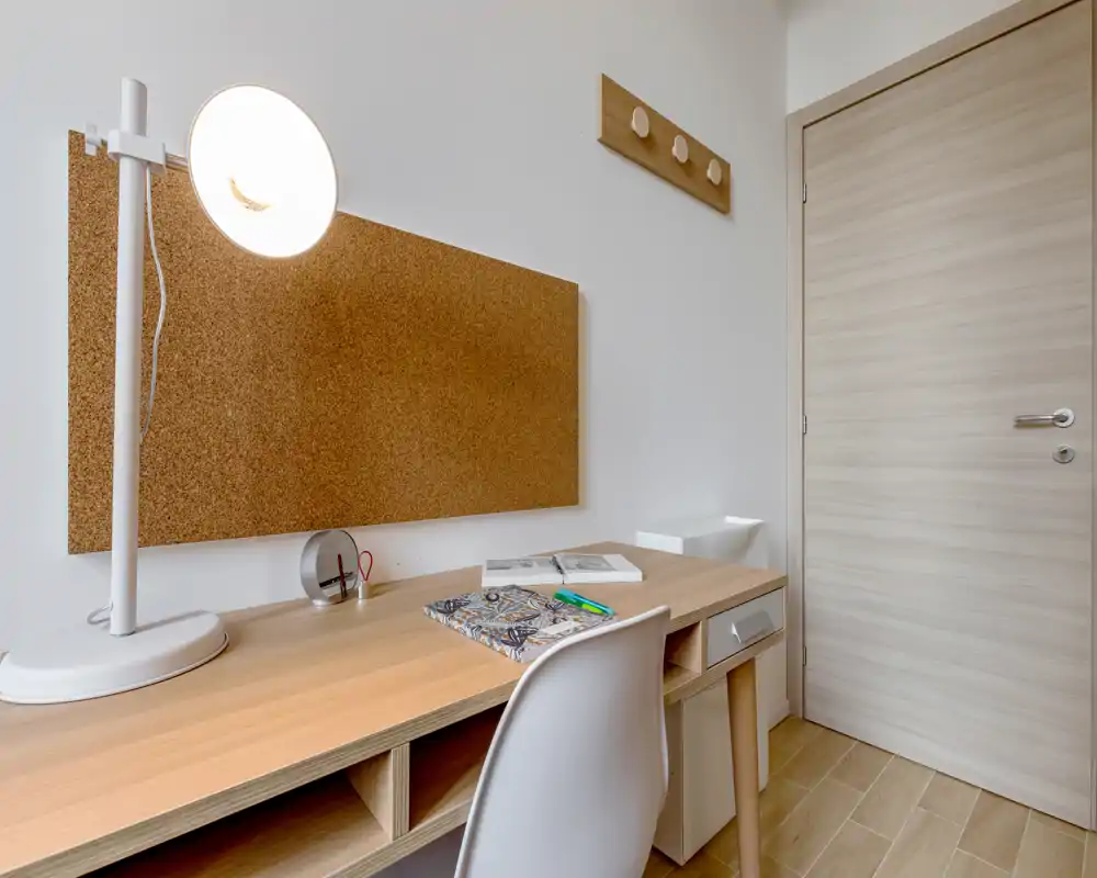 Área de escritorio luminosa y minimalista con panel de corcho, lámpara blanca y silla moderna — ideal para estudiar o trabajar en un apartamento compacto.