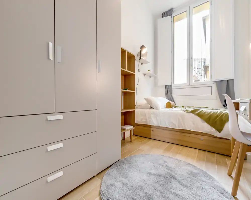 Dormitorio individual moderno y luminoso con cama arreglada, armario empotrado, escritorio y suelo de madera — ideal para estudiante o persona sola.