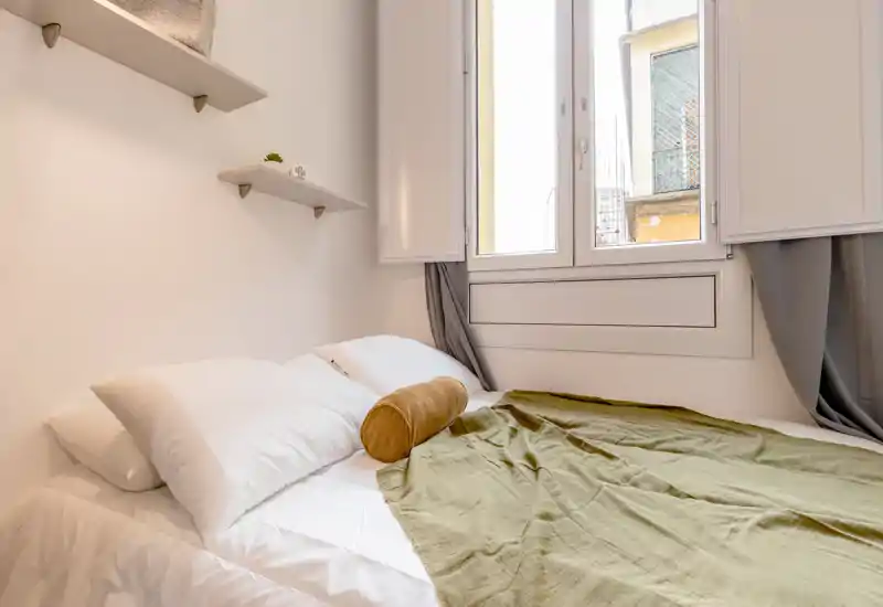 Dormitorio luminoso y compacto con cama bien hecha bajo una ventana grande, estantes flotantes y textiles cálidos que crean un ambiente acogedor.