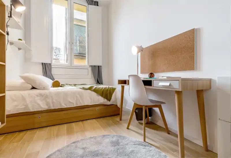 Dormitorio luminoso y compacto con cama individual, escritorio de madera y luz natural procedente de una gran ventana — ideal para estudiante o persona sola.
