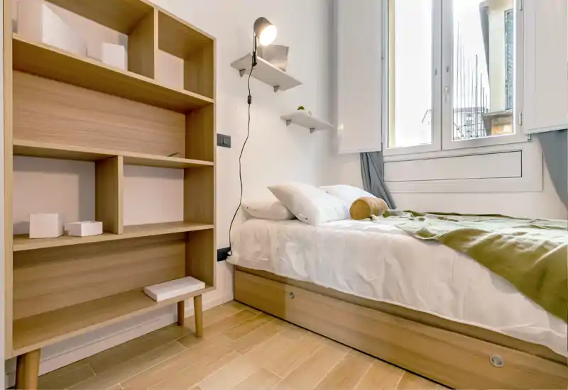 Dormitorio pequeño y luminoso con cama individual bien hecha, base de almacenamiento de madera y estantería abierta. La ventana grande proporciona luz natural y la decoración neutra crea un ambiente sereno.