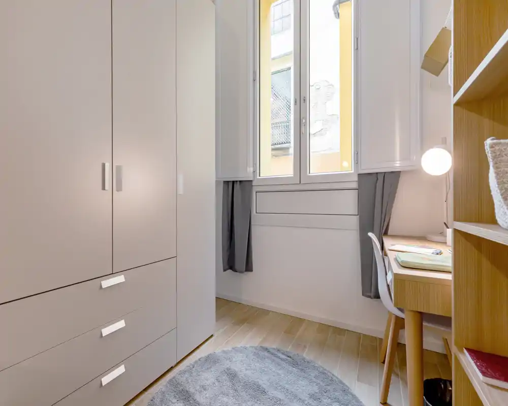 Espacio tipo dormitorio compacto con armario alto, escritorio y luz natural desde la ventana central — ordenado y funcional para una persona.