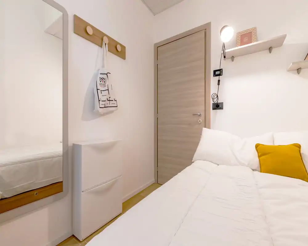 Dormitorio compacto y luminoso con una cama doble cómoda vestida en blanco y un cojín mostaza de contraste. Decoración minimalista y cuidada con lámpara de pared, estante, espejo y percheros — ideal para alquileres de corta estancia.