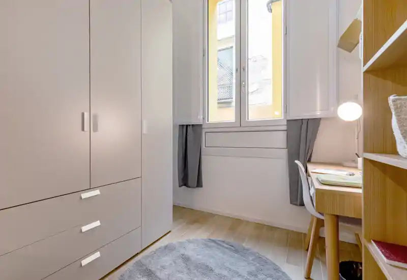 Compact bedroom-style space with a tall wardrobe, study desk and natural light from a central window — tidy and functional for a single occupant.