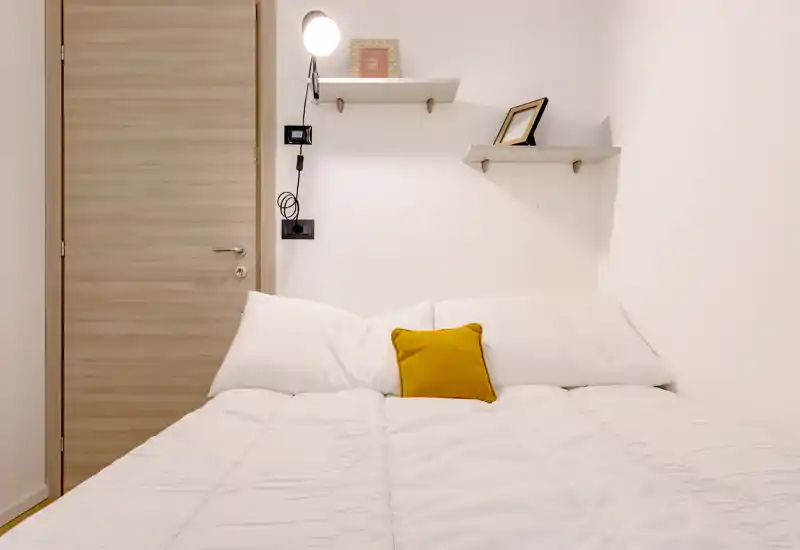 Dormitorio luminoso y minimalista con cama doble bien hecha, ropa de cama blanca y cojín amarillo; estantes y lámpara de pared crean un ambiente acogedor y moderno.
