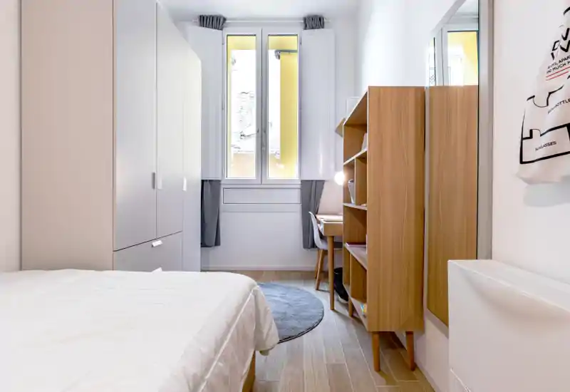 Dormitorio moderno y luminoso con cama bien hecha, estanterías de madera y un pequeño escritorio junto a la ventana. La luz natural y las líneas limpias crean un espacio tranquilo y acogedor.