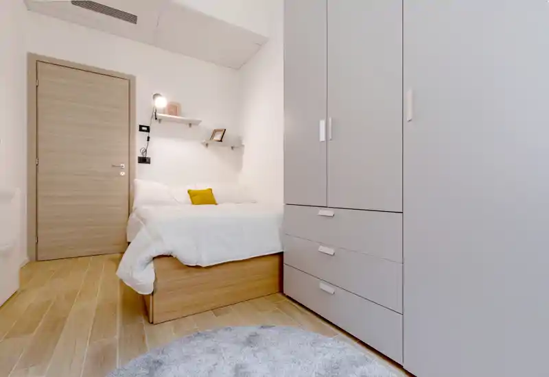 Dormitorio compacto y luminoso con cama plataforma de madera, ropa blanca y un cojín amarillo; armario empotrado y alfombra redonda que generan un ambiente acogedor.