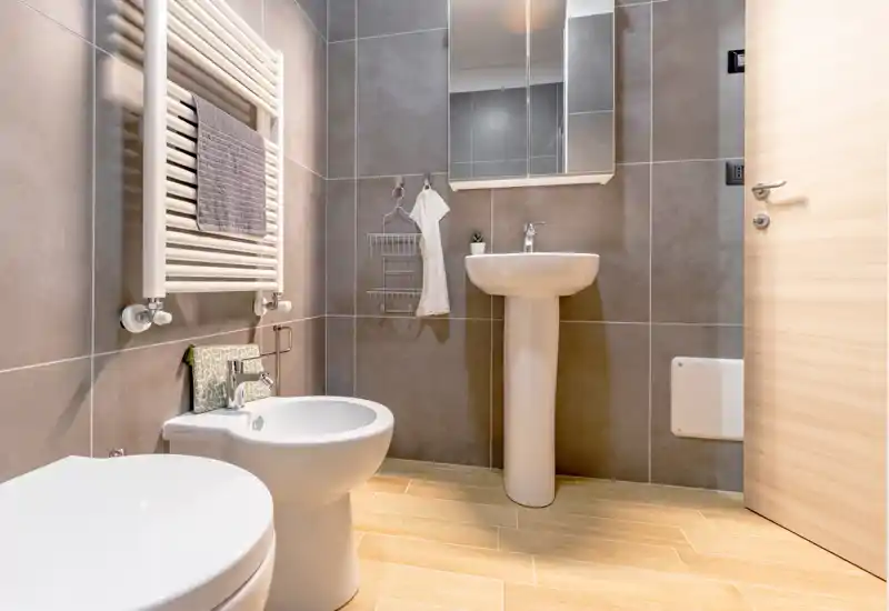 Modern, well-lit bathroom with pedestal sink, bidet and toilet, neutral gray tiles and warm wood-effect floor tiles. Compact, clean layout ideal for a rental property.