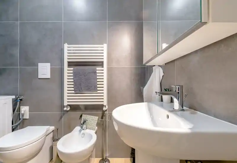 Modern, compact bathroom with a wall-mounted towel radiator, bidet, toilet and a white pedestal sink; neutral grey tiles and bright, clean styling.