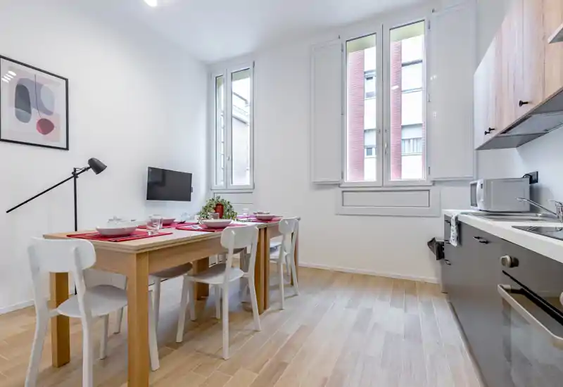 Bright, modern kitchen with a wooden dining table set for four, white chairs, and a compact kitchenette with appliances. Large windows provide abundant natural light and the styled table and plant add a welcoming touch.