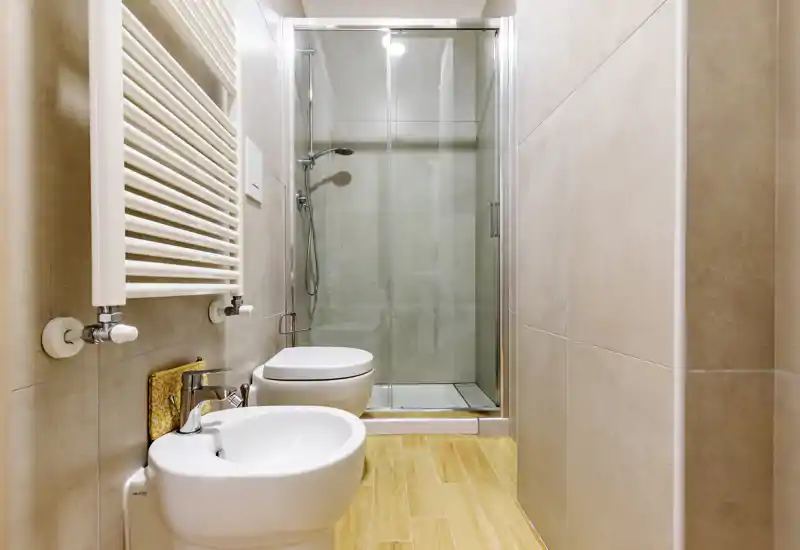 Modern compact bathroom with a walk-in glass shower, wall-mounted toilet, bidet and heated towel radiator; neutral tiles and wood-effect floor create a clean, contemporary look.