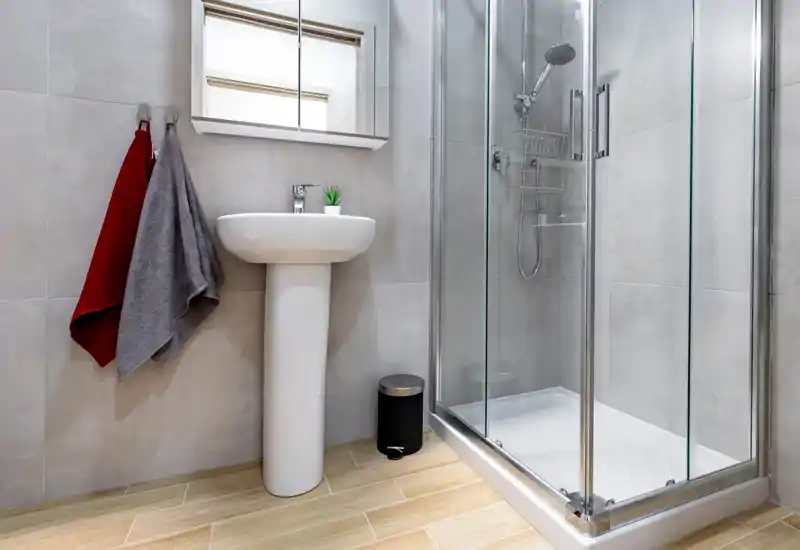 Bright, modern bathroom with a glass shower enclosure, pedestal sink, mirrored cabinet and wood-effect tile floor — clean, contemporary and well lit.