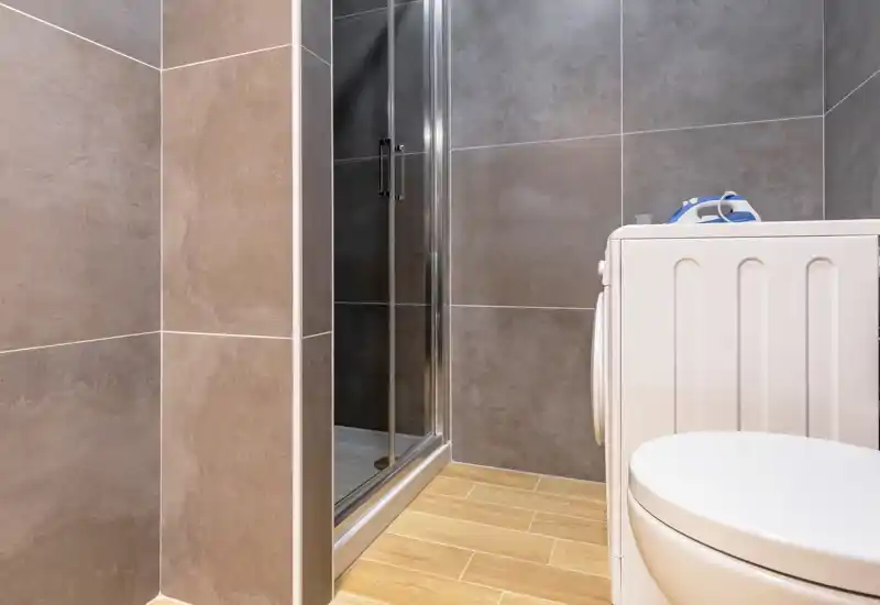Modern bathroom corner with a glass shower enclosure, wall tiles and a washing machine visible beside the toilet. Clean, contemporary finishes and warm floor tiles create a neat, functional space.