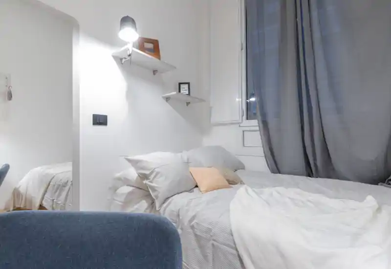 Dormitorio moderno y luminoso con cama doble bien hecha, almohadas apiladas y textiles suaves. La paleta neutra y la lámpara crean un ambiente tranquilo y acogedor, ideal para el anuncio.