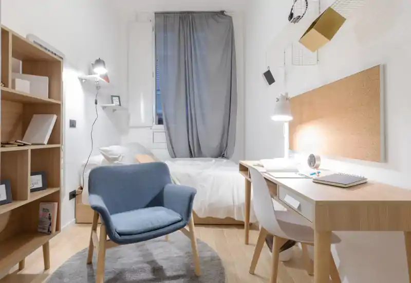 Dormitorio acogedor y luminoso con cama individual frente a cortinas grises, escritorio de madera con lámpara y un sillón cómodo, perfecto para estudiar o relajarse.