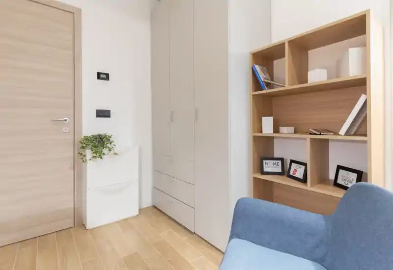 Pequeño rincón de estar luminoso con un sillón azul, armario empotrado y estantería de madera: decoración minimalista y una planta que aporta calidez.