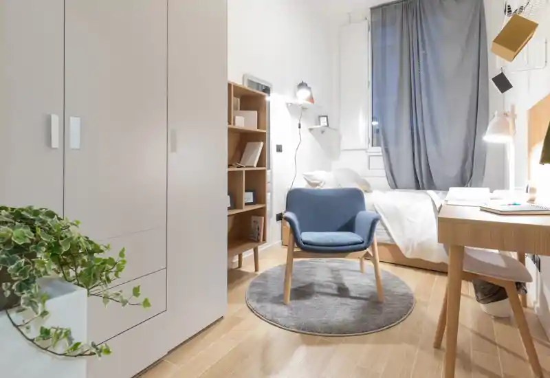 Dormitorio individual luminoso y bien decorado con cama cómoda, escritorio, armario y un sillón acogedor sobre una alfombra circular — ideal para estudiante o persona sola.