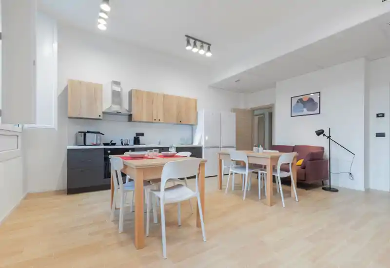 Bright, modern open-plan living area with a kitchenette, two dining tables and seating; clean wooden floors, a cozy armchair and contemporary lighting create a welcoming space.