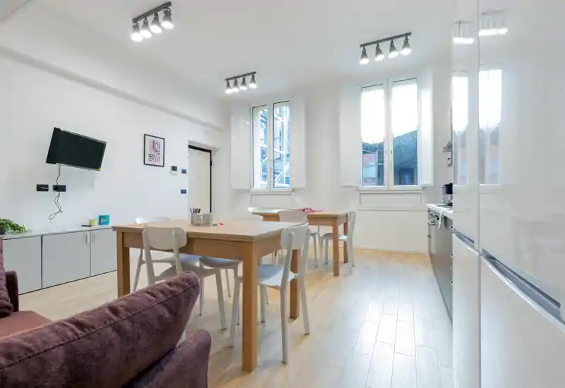 Bright, modern living/dining area with wooden tables, white chairs and a cozy sofa; large windows bring abundant natural light and a clean, minimalist style ideal for short-term rentals.