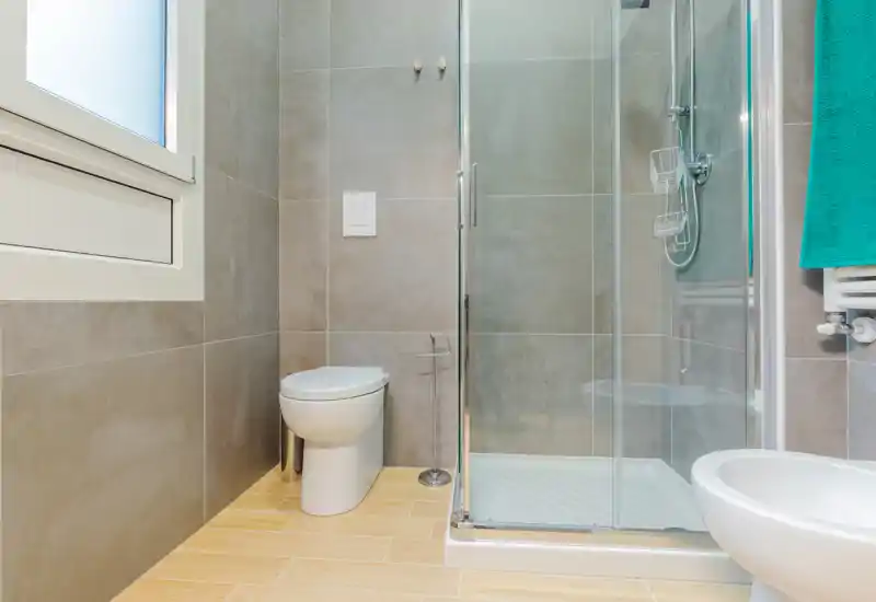 Modern, well-lit bathroom with a glass shower enclosure, wall-mounted toilet flush plate, and tiled walls and floor — clean and contemporary design.