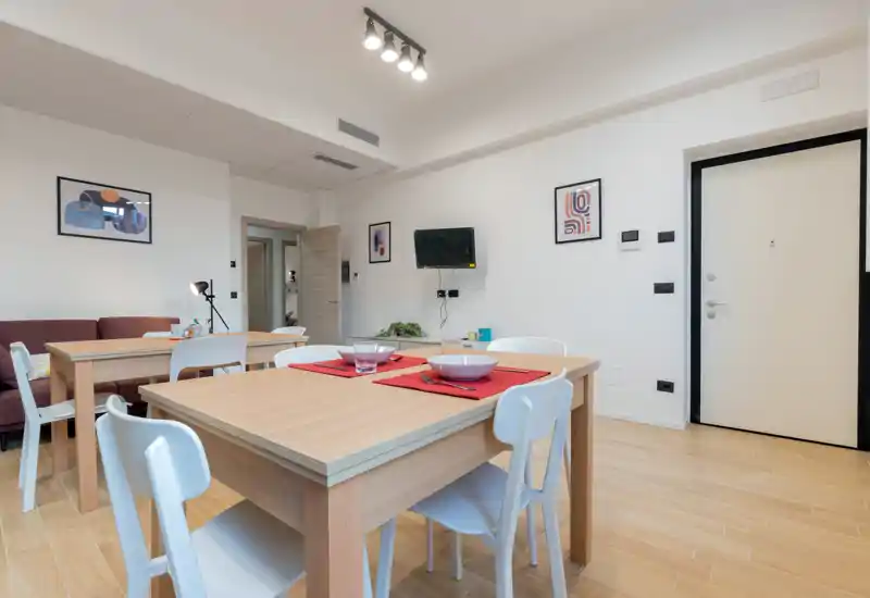 Bright, modern living/dining area with light wood tables, white chairs and a compact sofa; simple framed artwork and neat table settings create a welcoming, functional space.