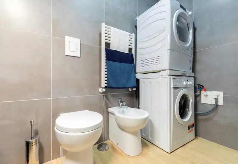 Compact modern bathroom with wall-mounted towel radiator, toilet, bidet and a stacked washer-dryer unit; light wood-effect floor and neutral grey tiles create a clean, contemporary look.