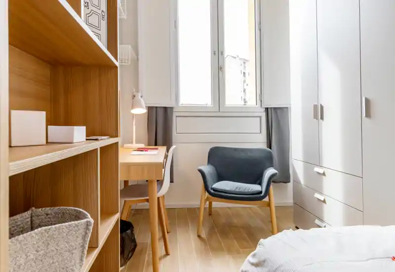 Chambre petite et lumineuse au style scandinave : étagères et sol en bois clair, petit bureau près de la fenêtre, fauteuil confortable et armoire. La lumière naturelle inonde la pièce, créant une atmosphère propre et accueillante, idéale pour la location.