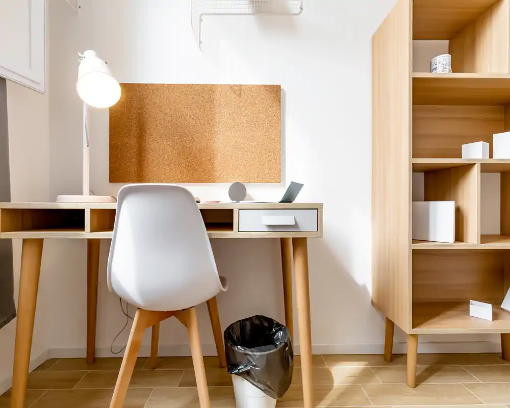 Angolo ufficio luminoso e minimale con scrivania in legno, sedia bianca, lampada da tavolo e pannello in sughero — uno spazio ordinato perfetto per lavoro o studio.