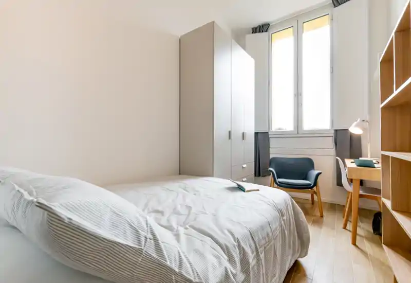 Camera da letto luminosa e ordinata con un letto ben visibile, armadio, poltrona per la lettura e una scrivania vicino alla finestra — ideale per una persona o per gli ospiti.