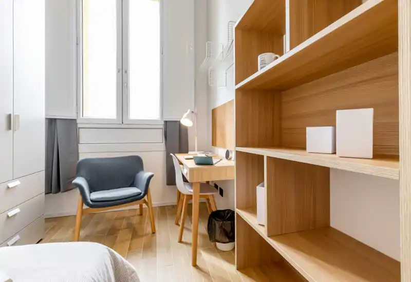 Camera da letto compatta e luminosa con poltrona accogliente, scrivania e scaffalature in legno; le tonalità calde e la luce naturale rendono l'ambiente ideale per una persona sola.
