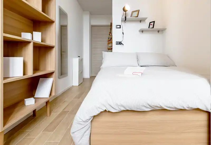 Camera da letto luminosa e minimal con letto matrimoniale ben rifatto su struttura in legno chiaro e scaffalatura coordinata; biancheria neutra e mensole a parete creano un’atmosfera moderna e tranquilla.