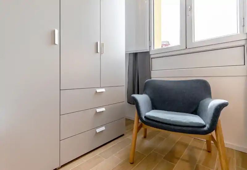 Bright, minimal corner with a modern armchair beside a built-in wardrobe and a large window — ideal for a reading nook or compact living area.