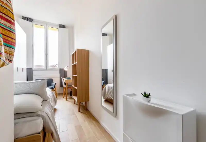 Camera da letto luminosa e minimal con letto ben rifatto, una grande finestra che illumina la stanza, una scrivania piccola e una libreria in legno.