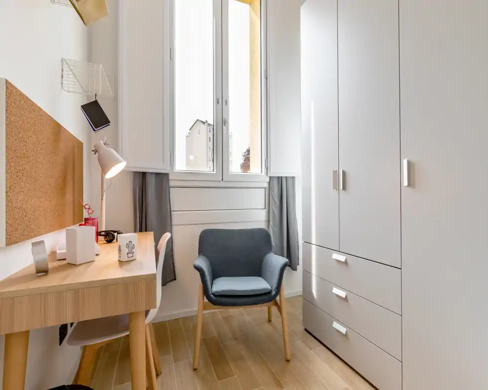 Rincón de dormitorio compacto y bien iluminado con escritorio, butaca y gran armario; la luz natural de la ventana realza la decoración neutra de estilo escandinavo.