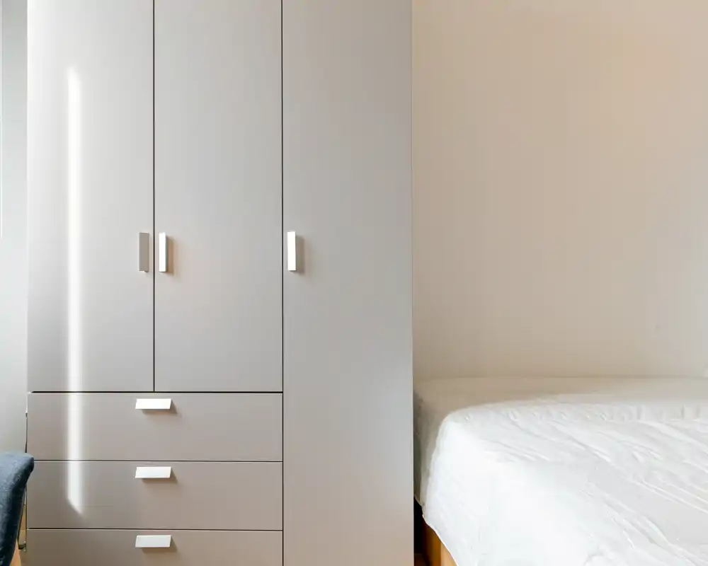 Habitación luminosa y minimalista con un armario y el borde de una cama con ropa blanca. Líneas limpias y luz natural crean un ambiente tranquilo y ordenado, perfecto para anuncios de alquiler.