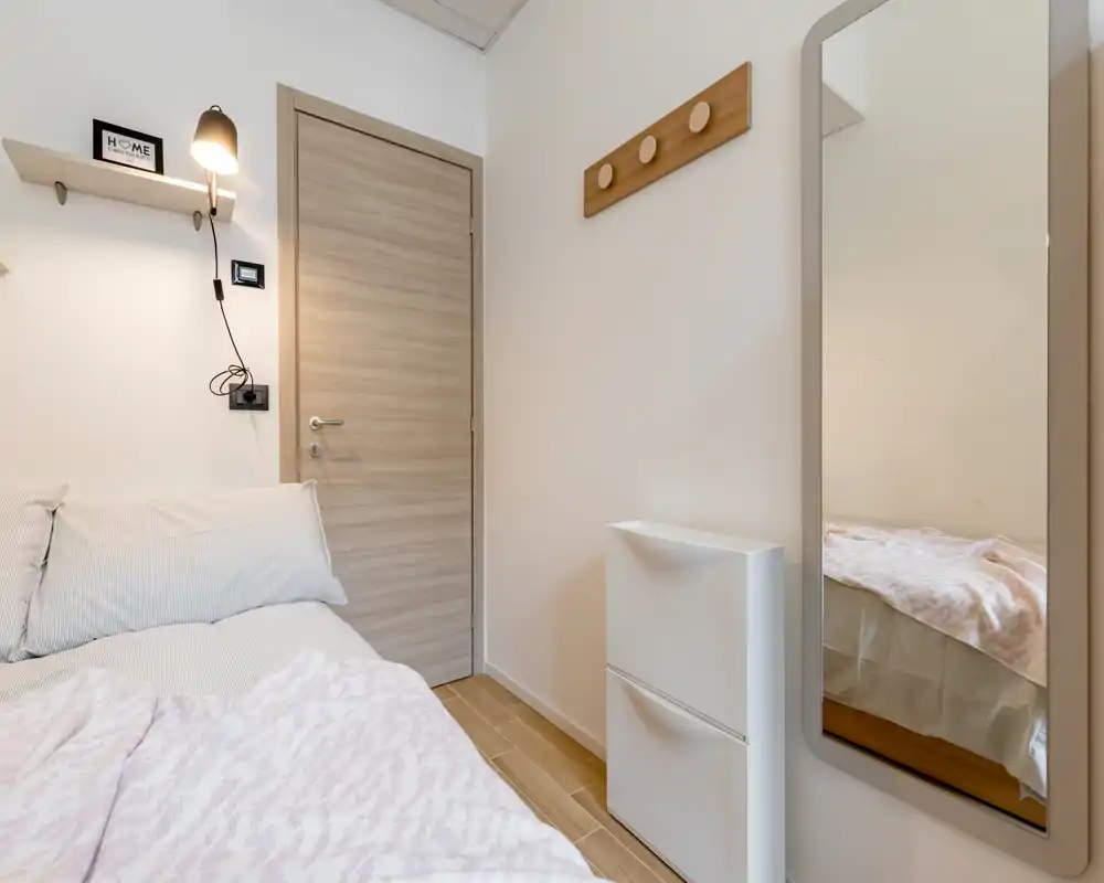 Compact, softly lit bedroom corner with a neatly made bed, wall lamp, coat hooks and a full-length mirror reflecting part of the mattress. Neutral tones and wood flooring give a calm, modern feel.