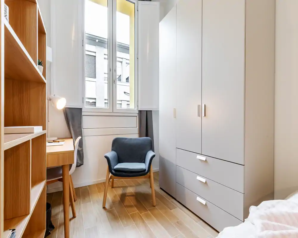 Compact, well-lit bedroom corner with a soft armchair, built-in wardrobe and a partial view of the bed. Clean Scandinavian-style wood shelving and desk make this corner ideal for working or relaxing.