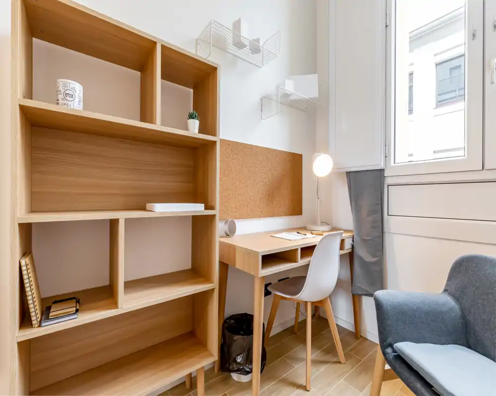 Bright, compact study corner with a wooden desk, shelving and a comfortable armchair next to a large window — ideal for working or reading.