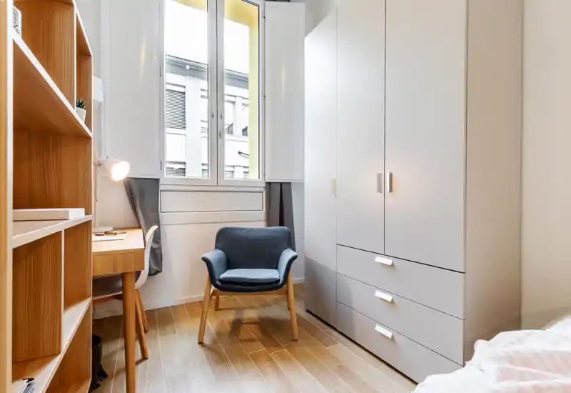 Small, bright bedroom corner with a wardrobe, armchair and a study desk by the window. The space is modern and well lit, ideal for a single occupant.