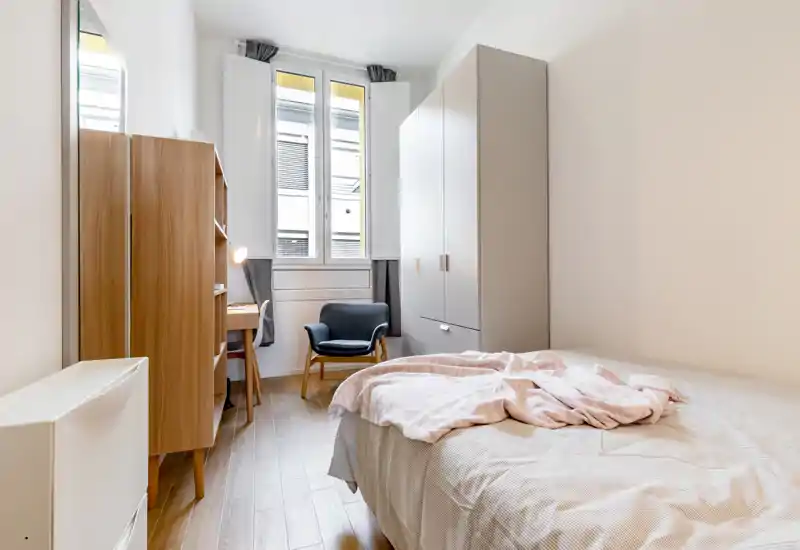 Bright, compact bedroom with a comfortable double bed, wardrobe, desk and armchair by the window. Neutral tones and natural light make it airy and welcoming — ideal for a single occupant or couple.