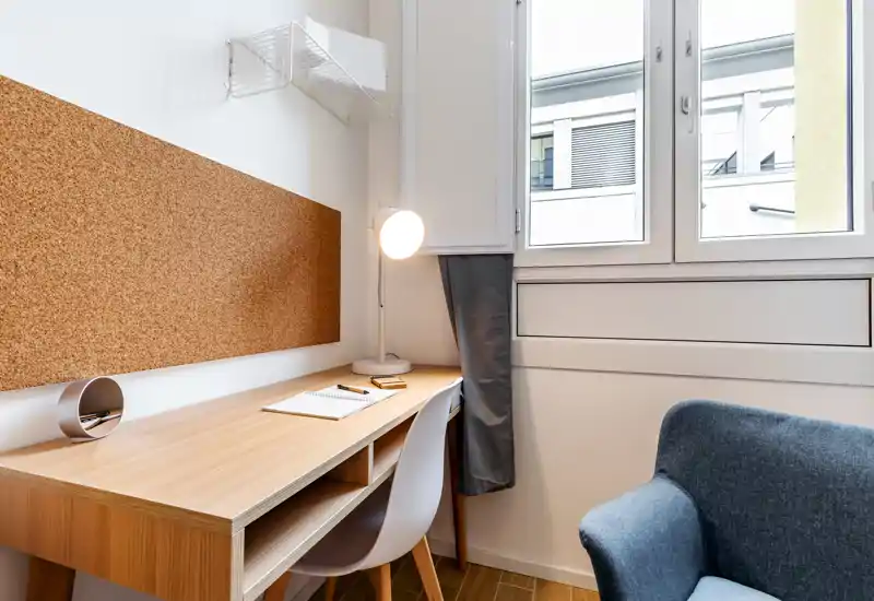 Bright, compact living area with a wooden desk, cork noticeboard and a cozy armchair by the window — ideal as a home-office corner.