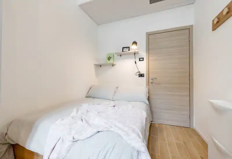 Compact, well-lit bedroom with a double bed, soft linens and a cozy throw. Neutral tones and simple shelving create a calm, modern look ideal for short-term rentals.