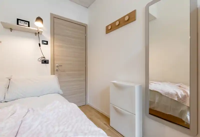 Compact, softly lit bedroom corner with a neatly made bed, wall lamp, coat hooks and a full-length mirror reflecting part of the mattress. Neutral tones and wood flooring give a calm, modern feel.