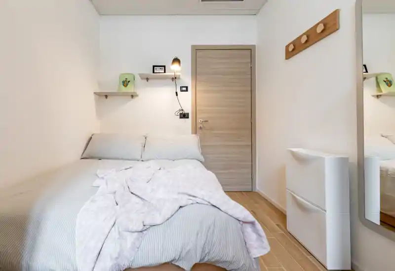 Bright, compact bedroom with a neatly made double bed, soft linens and a cozy throw. Minimal Scandinavian-style décor, wall shelves and a bedside lamp create a calm, functional sleeping space.