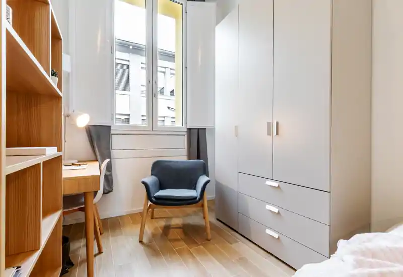 Compact, well-lit bedroom corner with a soft armchair, built-in wardrobe and a partial view of the bed. Clean Scandinavian-style wood shelving and desk make this corner ideal for working or relaxing.